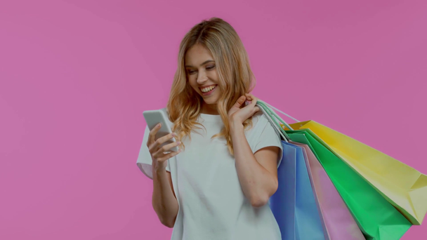 Excited woman using smartphone and holding shopping bags isolated on purple - Felvétel, videó