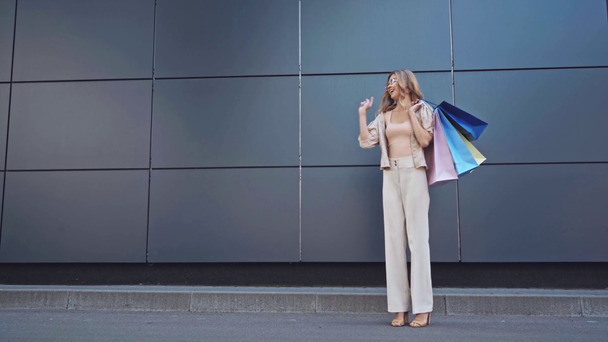 Blonde Frau zeigt mit Finger auf Einkaufstüten in der Nähe von Gebäude  - Filmmaterial, Video