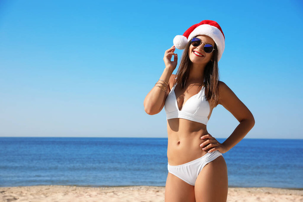 Young woman wearing Santa hat and bikini on beach, space for text. Christmas vacation - Foto, Imagen