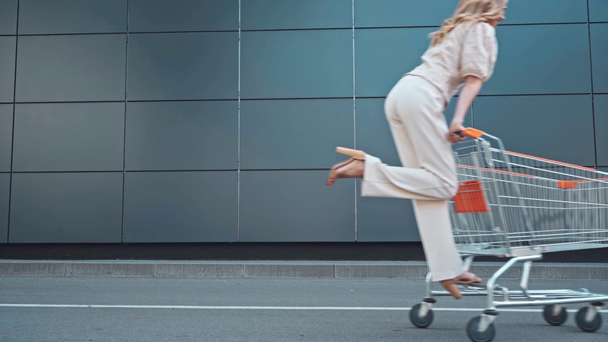 Blonde woman riding on shopping cart outdoors  - Footage, Video