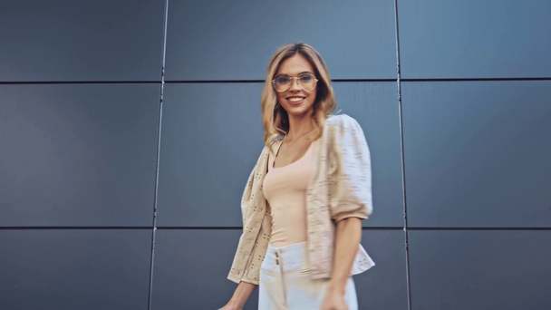 Блондинка в солнечных очках держит сумки возле здания  - Кадры, видео