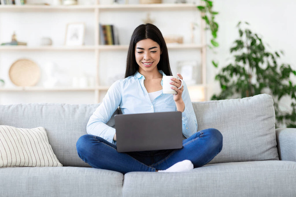 Concepto de ocio. Chica asiática relajarse en el sofá, beber café y usar el ordenador portátil - Foto, imagen