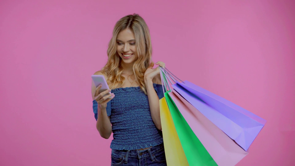 Blondýny žena s nákupními taškami pomocí smartphone izolované na fialové - Záběry, video