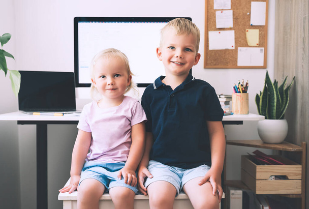 Portrait of preschool boy and girl on background of computer with blank white screen in home interior. Kids and computer technologies. Children online learning, development or entertainment. - Photo, image