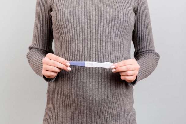 Gesneden beeld van positieve zwangerschapstest met twee strepen tegen de buik van zwangere vrouw op grijze achtergrond. Toekomstige moeder in grijze jurk. Zwangerschap verrassing. Kopieerruimte. - Foto, afbeelding