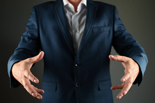 Ένας άντρας με κοστούμι μιμείται το να κρατάει ένα αντικείμενο. Ένας κουστουμάτος απλώνει τα χέρια του σε μαύρο φόντο. Έννοια: ένα θετικό μήνυμα στις επιχειρήσεις - Φωτογραφία, εικόνα