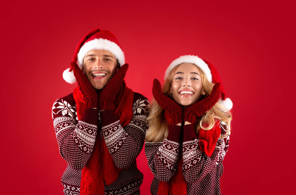 Es ist Weihnachtszeit. Entzückendes junges Paar in Fäustlingen, Nikolausmützen und Schals vor rotem Hintergrund - Foto, Bild