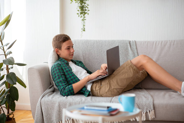 Side view of teen boy lying on couch, using laptop - Foto, afbeelding