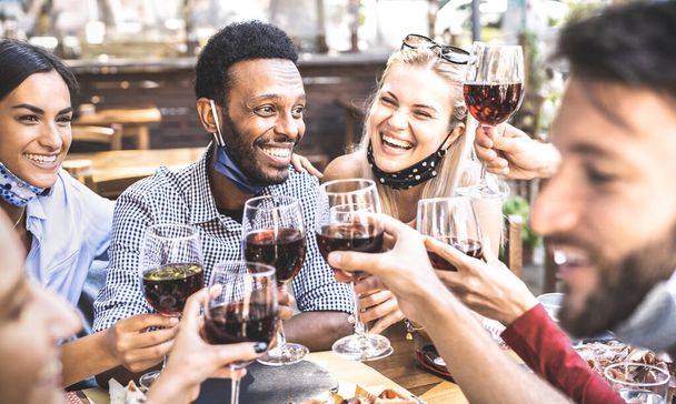 Friends toasting red wine at outdoor restaurant bar with open face mask - New normal lifestyle concept with happy people having fun together on warm filter - Focus on afroamerican guy - Photo, Image