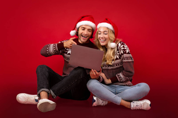 Full length portrait of young couple in Christmas outfits pointing at laptop computer on red background - Fotoğraf, Görsel