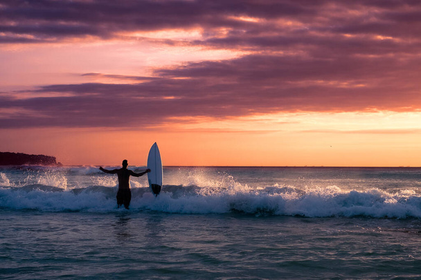 Challenge and conquer the concept: a silhouette of a surfer holding a Board and looking at the ocean against the sunset background - 写真・画像
