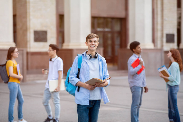 Студент, стоящий с книгой и рюкзаком, позирует возле здания колледжа - Фото, изображение