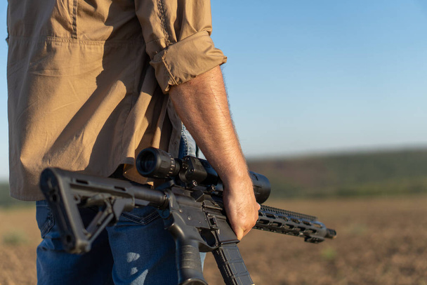 Винтовка в руках человека, крупным планом. Охотник с оружием на охоте на фоне красивого летнего пейзажа - Фото, изображение
