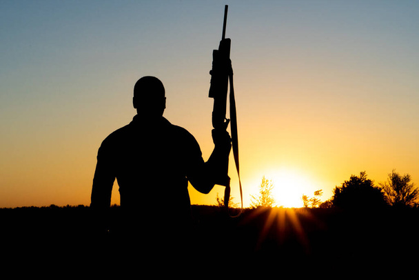 Silueta de un hombre con un arma en sus manos sobre un fondo al atardecer - Foto, Imagen