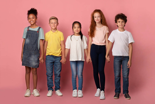 Schulfreundschaft. Entzückende multiethnische Kinder, die sich auf rosa Hintergrund die Hände reichen - Foto, Bild