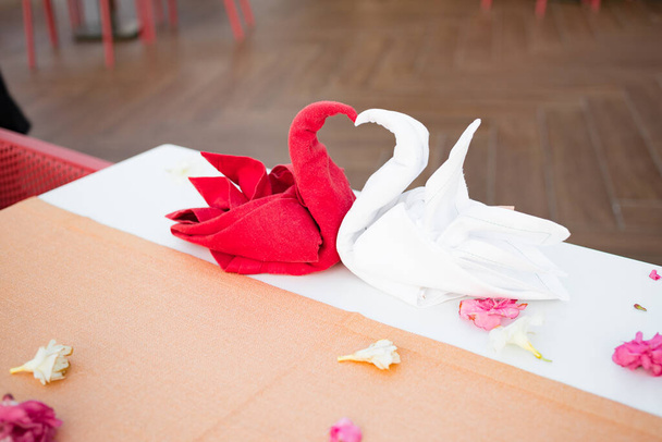 Serviette pliée rouge et blanc en forme d'oiseau de cygne dans un restaurant sur une table blanche dans un resort. Une fleur pétales et verres sur la table. Dîner romantique pour les jeunes mariés. Coucher de soleil. - Photo, image
