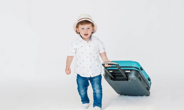A boy with glasses and a hat is going to go on vacation, looks slyly from under his glasses. Leaning on a blue suitcase. The child is happy, smiling and laughing. Summer, vacation. - Fotografie, Obrázek