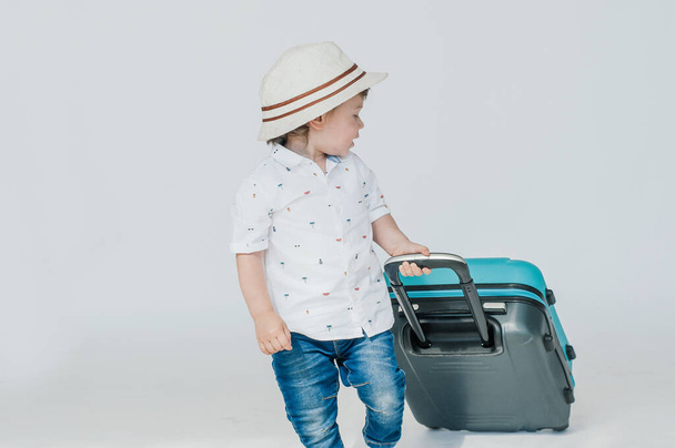 A boy with glasses and a hat is going to go on vacation, looks slyly from under his glasses. Leaning on a blue suitcase. The child is happy, smiling and laughing. Summer, vacation. - Φωτογραφία, εικόνα