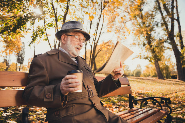 Profile side photo of excited old man pensioner rest relax fall park outside have break pause hold takeout coffee beverage mug read incredible news paper wear coat jacket cap headwear - Photo, image