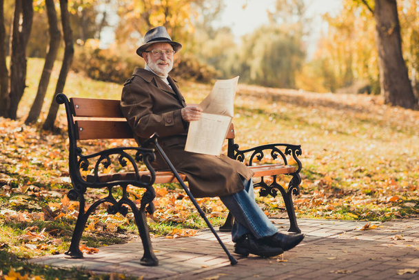 Hello sunny weather day autumn concept. Full length photo of positive old man pensioner enjoy park forest relax sit bench hold read newspaper wear season outerwear cap - Zdjęcie, obraz