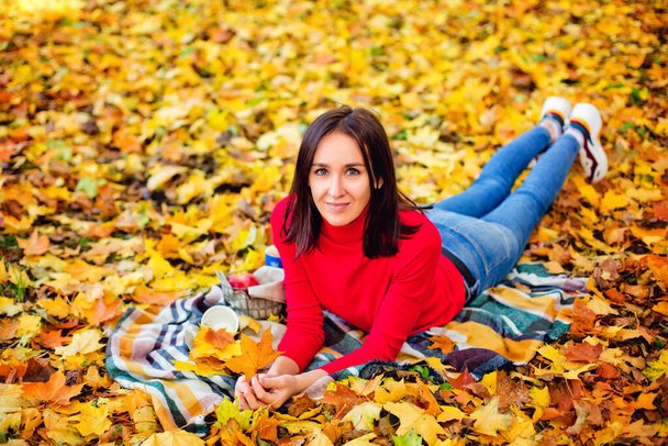 Ciemnowłosa kobieta w czerwonym swetrze leży na kocu w parku pośród żółtych liści klonu. Jesienny piknik w przyrodzie. - Zdjęcie, obraz