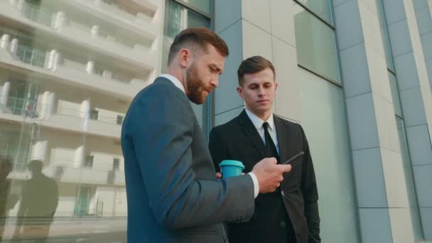 Dos empleados masculinos interactuando entre sí en el distrito de negocios del centro. Empresarios confiados que usan teléfonos inteligentes discutiendo la planificación de proyectos de negocios de computadoras en equipo corporativo - Metraje, vídeo