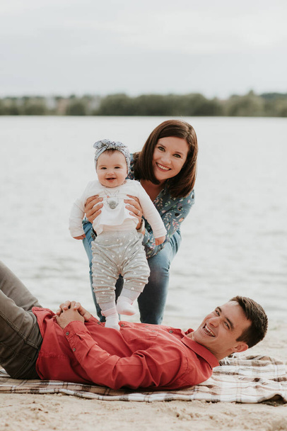 Happy young family sitting near the water. Mom, dad and their little baby daughter sitting together. Happy family outdoors. Smiling people - Foto, Imagem