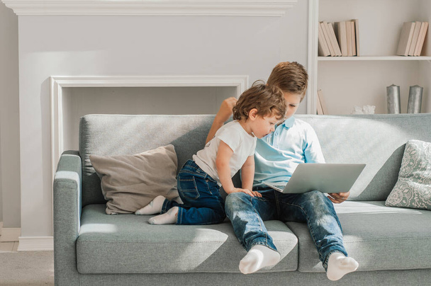 Two little kids siblings seated on sofa in living room holding on lap laptop play online game, watch cartoons spend weekend using electronic device, bad habit, generation addicted with gadgets concept - Foto, Imagem