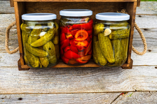 Glass jars with pickled red bell peppers and pickled cucumbers (pickles) isolated in wooden crate. Jars with variety of pickled vegetables. Preserved food concept in a rustic composition. - Photo, Image