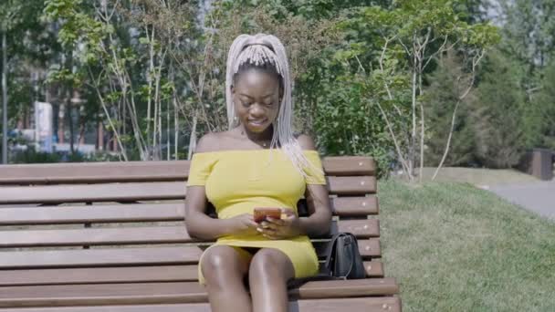 Stylowa kobieta z białymi warkoczami za pomocą telefonu komórkowego siedzi na ławce - Materiał filmowy, wideo