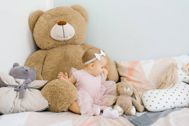 A cute one year old girl in a pink dress sits on the bed in the children's room at home among soft funny toys and looks at the Bunny - Photo, image