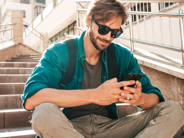 Handsome stylish smiling hipster lambersexual model.Man dressed in white T-shirt.Fashion male sitting on the stairs. Holding, scrolling and texting in his cellphone. Writes sms on smartphone - Photo, Image