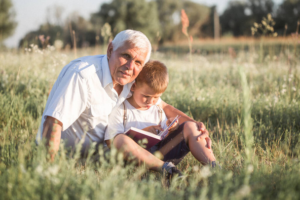 Mid shot of grandfather and his grandson while reading a book together in meadow Small boy making first steps in read. - Photo, Image