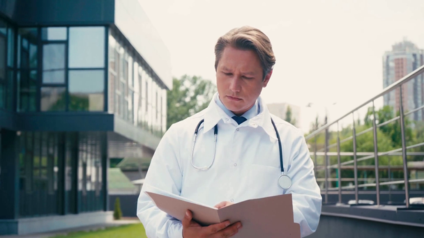 白いコートを着た重病の医者がフォルダを保持し外のクリニックの近くを歩く - 映像、動画