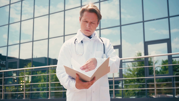 médico em casaco branco olhando para documentos na pasta perto da clínica  - Filmagem, Vídeo