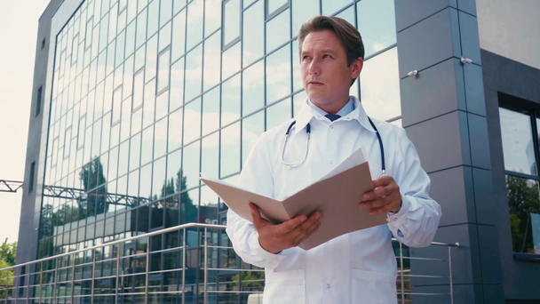 врач в белом халате и стетоскопе держит папку рядом с клиникой снаружи - Кадры, видео