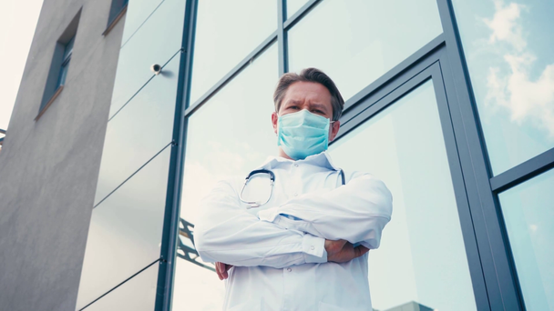 vista a basso angolo del medico in maschera medica in piedi con le braccia incrociate all'esterno - Filmati, video