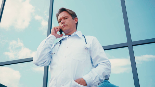 низький кут зору лікаря стоїть з рукою в кишені і розмовляє на смартфоні
 - Кадри, відео