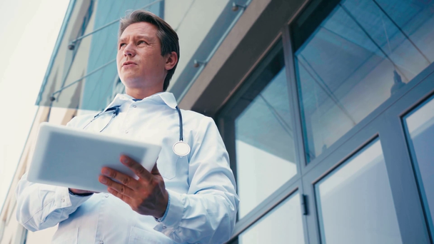 vista a basso angolo del medico in camice bianco utilizzando tablet digitale e parlando all'esterno  - Filmati, video