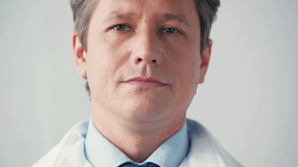 vážný lékař v bílém plášti při pohledu na fotoaparát izolovaný na šedé - Záběry, video