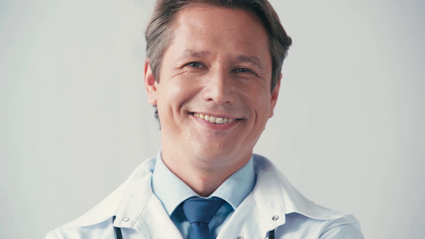 vrolijke dokter in witte jas kijkend naar camera op grijs - Video