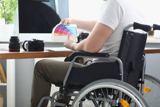Man in white T-shirt and green pant sit in wheelchair and hold multi-colored palette in his hand. - Foto, Imagen