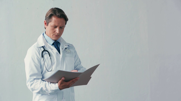 serious doctor in white coat holding and looking at folder on grey - Footage, Video