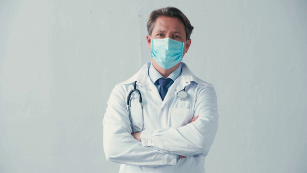 Beyaz önlüklü ve tıbbi maskeli doktor gri kollarıyla ayakta duruyor. - Video, Çekim