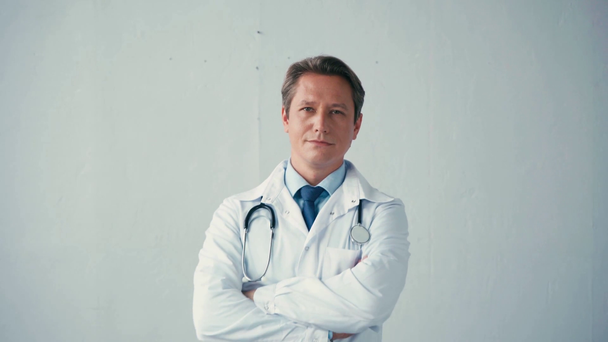 confident doctor in white coat standing with crossed arms on grey  - Video, Çekim