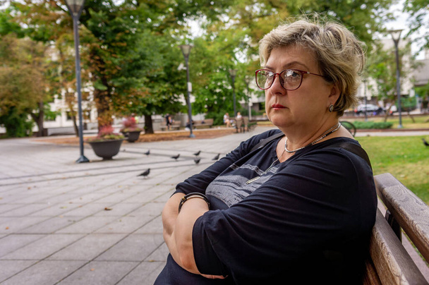 Kypsä surullinen nainen mustassa mekossa ja lasit katsoo kaukaisuuteen. Lepää kaupungin puistossa - Valokuva, kuva