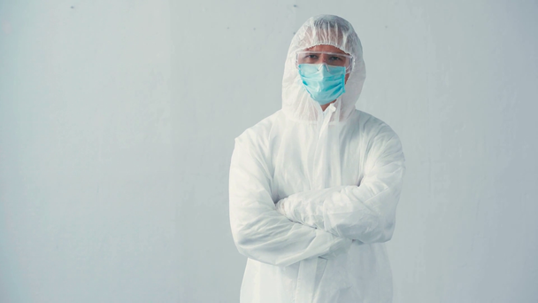 científico en traje de materiales peligrosos y máscara médica de pie con los brazos cruzados en gris - Metraje, vídeo