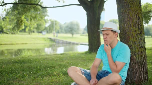 Nuori hattupäinen mies istuu järven rannalla puun alla, kopioi tilaa, kameran liikettä - Materiaali, video