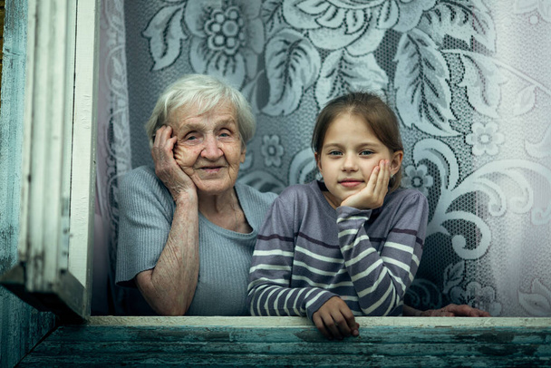 Μια ηλικιωμένη γυναίκα με μια μικρή εγγονή μαζί κοιτούν έξω από το παράθυρο του σπιτιού.. - Φωτογραφία, εικόνα