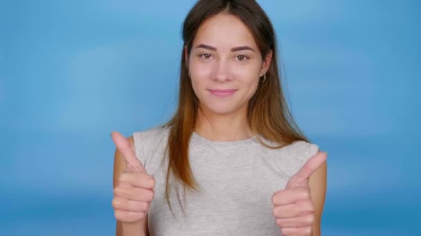 Gri tişörtlü güzel, pozitif kadın parmaklarını yukarı kaldırıyor ve mavi arka planda fotokopi çekerek gülümsüyor. 4K yavaş çekim görüntüleri - Video, Çekim